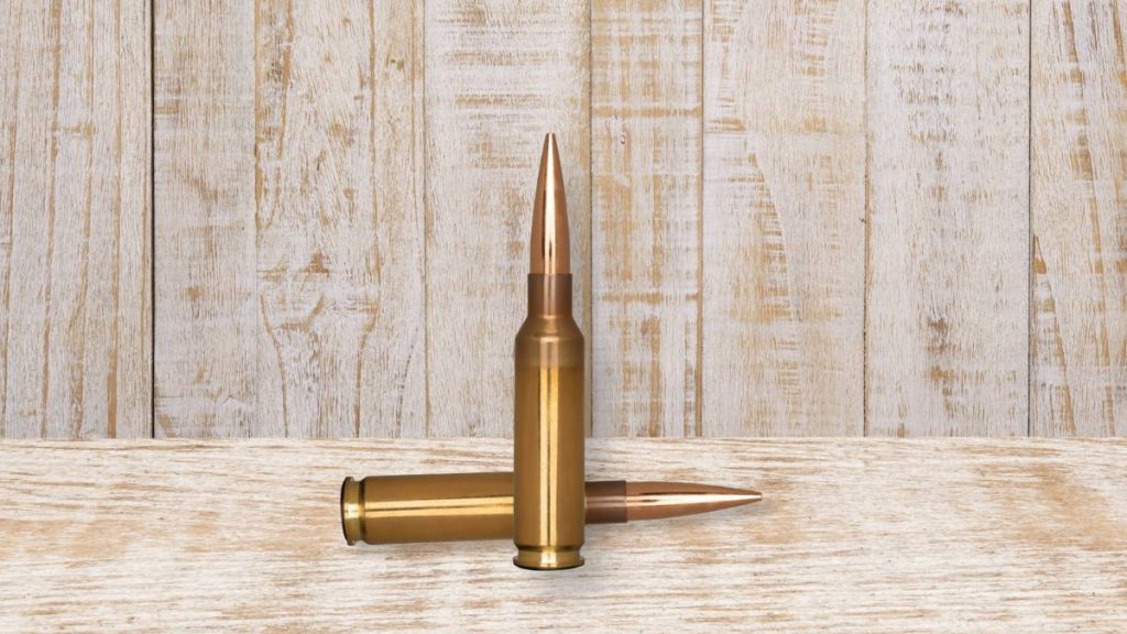 There are two 6.5 Creedmoor cartridges, in which one standing upright and the other lying down, on a wooden table.