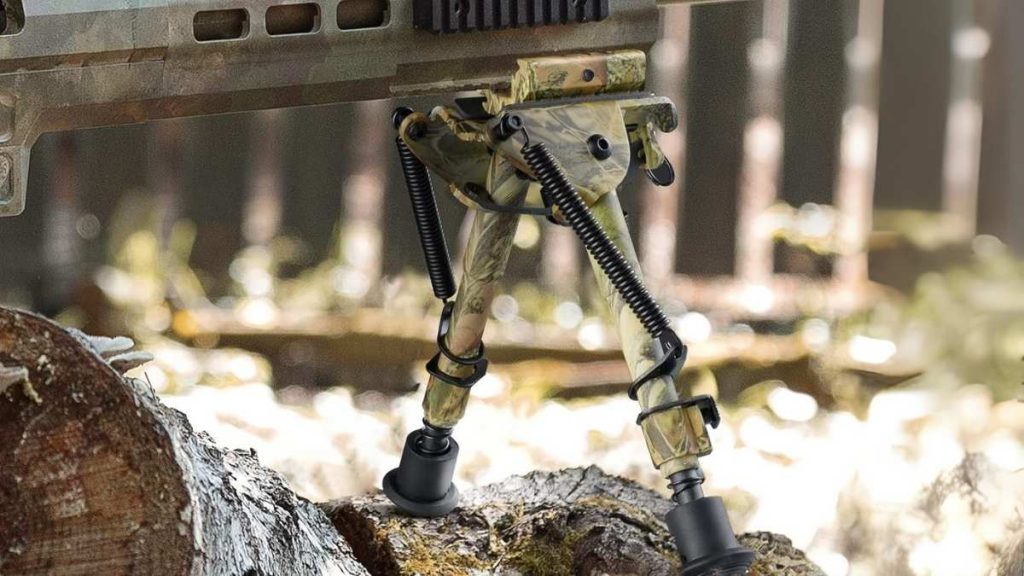 A camouflaged military bipod stands on a log, showcasing its adjustable legs in a forested background.